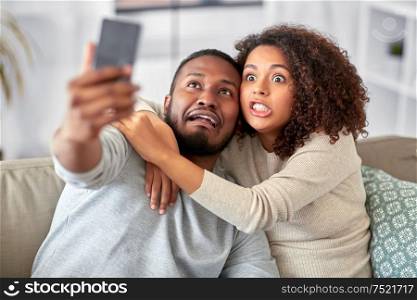 family, technology and people concept - african american couple with smartphone taking selfie and making funny faces at home. couple with smartphone taking selfie at home