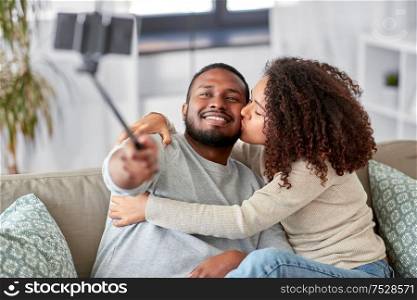 family, technology and people concept - happy african american couple taking picture by smartphone and selfie stick at home. couple taking picture by selfie stick at home