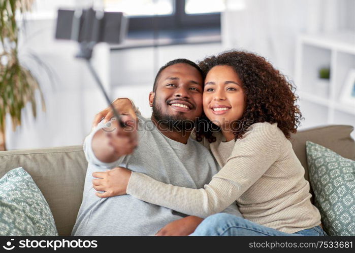 family, technology and people concept - happy african american couple taking picture by smartphone and selfie stick at home. couple taking picture by selfie stick at home