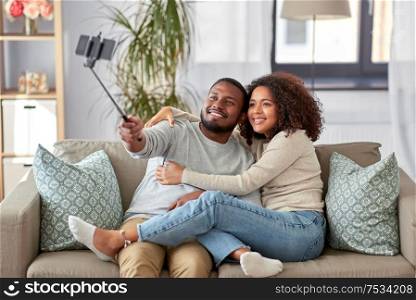 family, technology and people concept - happy african american couple taking picture by smartphone and selfie stick at home. couple taking picture by selfie stick at home