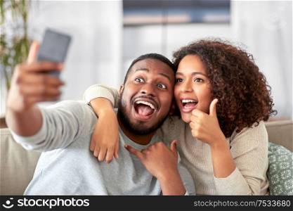 family, technology and people concept - happy african american couple with smartphone taking selfie and showing thumbs up and call gesture at home. happy couple with smartphone taking selfie at home