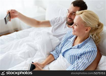 family, technology and people concept - happy couple with smartphone taking selfie in bed at home. happy couple with smartphone taking selfie at home