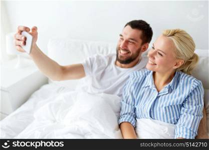 family, technology and people concept - happy couple with smartphone taking selfie in bed at home. happy couple with smartphone taking selfie at home
