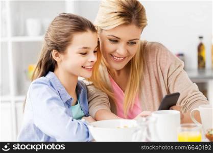 family, technology and people concept - happy mother and daughter with smartphone having breakfast at home kitchen. family with smartphone having breakfast at home