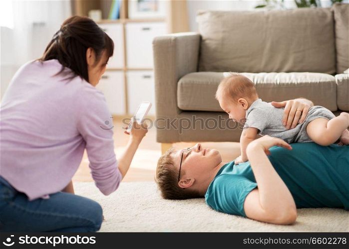 family, technology and people concept - happy mother and father taking picture or recording video of baby boy with smartphone at home. mother picturing father with baby by smartphone