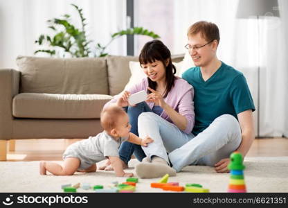 family, technology and people concept - happy mother and father taking picture or recording video of baby boy with smartphone at home. happy family with baby boy at home