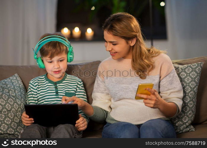 family, technology and people concept - happy mother and little son sitting on sofa with tablet pc computer, headphones and smartphone at home. mother and son using gadgets at home