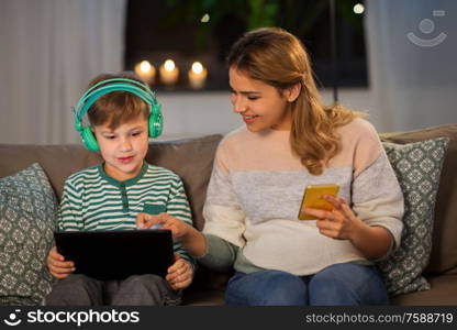 family, technology and people concept - happy mother and little son sitting on sofa with tablet pc computer, headphones and smartphone at home. mother and son using gadgets at home