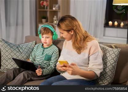 family, technology and people concept - happy mother and little son sitting on sofa with tablet pc computer, headphones and smartphone at home. mother and son using gadgets at home