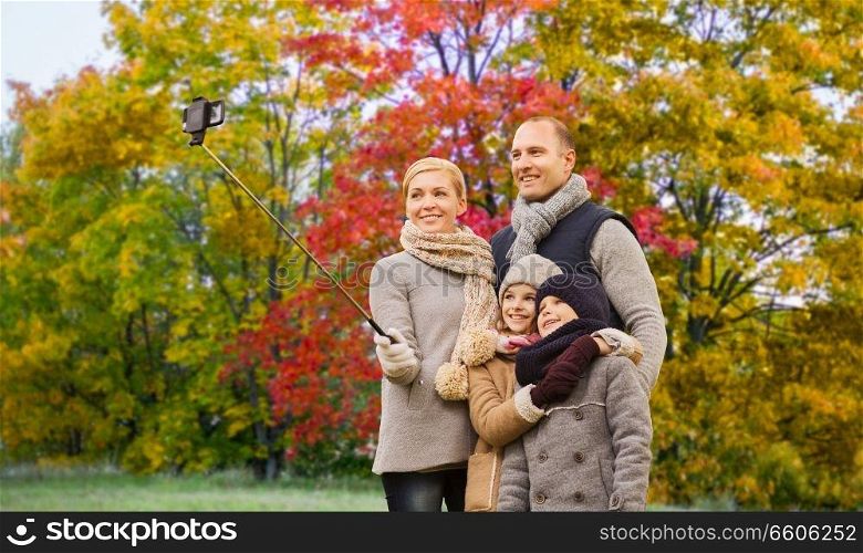 family, technology and people concept - happy mother, father, daughter and son taking picture by smartphone on selfie stick over autumn park background. family taking selfie over autumn park background