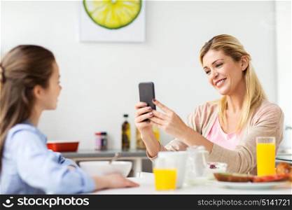 family, technology and people concept - happy mother with smartphone having breakfast and photographing her daughter at home kitchen. woman photographing daughter by smartphone at home