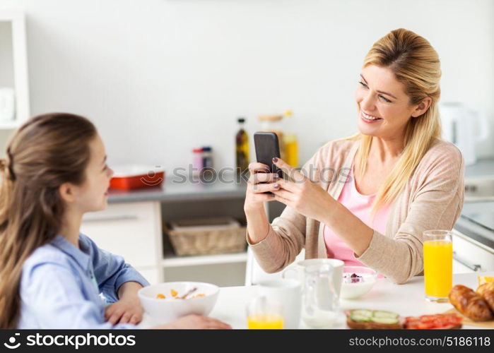 family, technology and people concept - happy mother with smartphone having breakfast and photographing her daughter at home kitchen. woman photographing daughter by smartphone at home