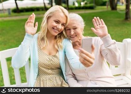 family, technology and people concept - happy smiling young daughter and senior mother with smartphone taking selfie and waving hands at park. daughter and senior mother taking selfie at park