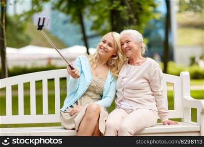 family, technology and people concept - happy smiling young daughter and senior mother sitting on park bench and taking picture with smartphone selfie stick. daughter and senior mother taking selfie at park