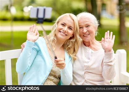 family, technology and people concept - happy smiling young daughter and senior mother sitting on park bench and taking picture with smartphone selfie stick. daughter and senior mother taking selfie at park