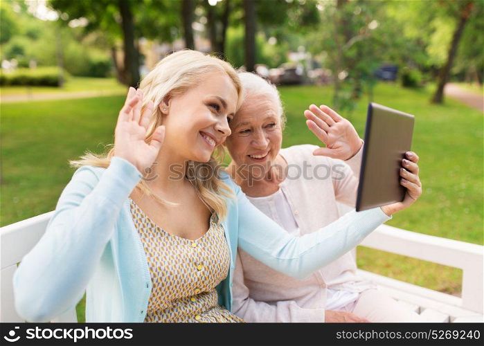 family, technology and people concept - happy smiling young daughter with tablet pc computer and senior mother on park bench waving hands. daughter with tablet pc and senior mother at park