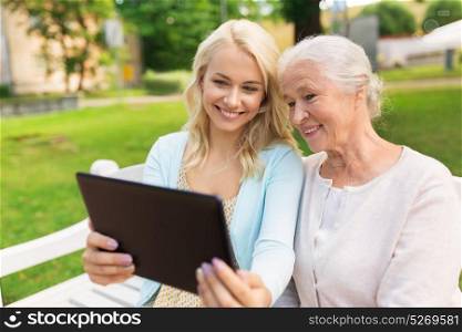 family, technology and people concept - happy smiling young daughter with tablet pc computer and senior mother sitting on park bench. daughter with tablet pc and senior mother at park