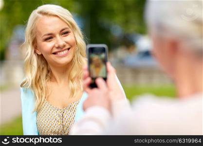 family, technology and people concept - senior mother with smartphone photographing her happy smiling young daughter at park. senior mother photographing daughter by smartphone