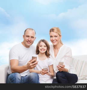 family, technology and people concept - smiling mother, father and little girl with smartphones over blue sky background