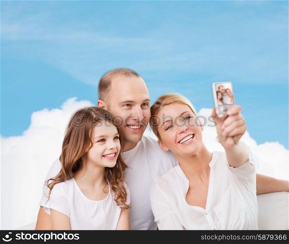 family, technology and people concept - smiling mother, father and little girl making selfie with camera over blue sky and white cloud background