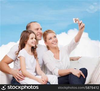 family, technology and people concept - smiling mother, father and little girl making selfie with camera over blue sky and white cloud background