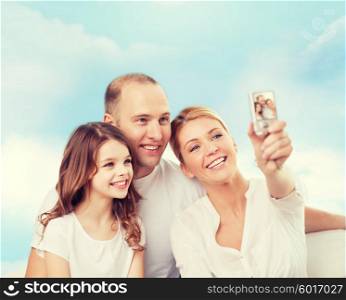 family, technology and people concept - smiling mother, father and little girl making selfie with camera over blue sky background