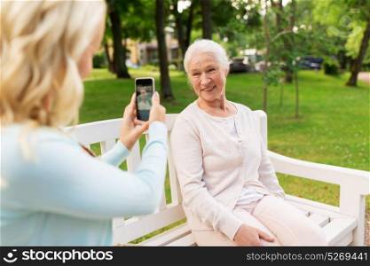 family, technology and people concept - young daughter with smartphone photographing her happy smiling senior mother at park. daughter photographing senior mother by smartphone
