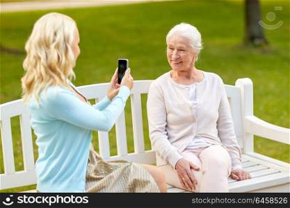 family, technology and people concept - young daughter with smartphone photographing her happy smiling senior mother at park. daughter photographing senior mother by smartphone