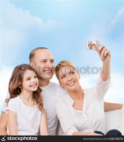family, technology and people - smiling mother, father and little girl making selfie with camera over blue sky background