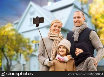 family, technology and real estate concept - happy mother, father, daughter and son taking picture by smartphone on selfie stick over living house background outdoors in autumn. family takes autumn selfie by cellphone over house