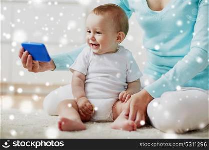 family, technology, child and parenthood concept - happy smiling young mother showing smartphone to little baby at home over snow. happy mother showing smartphone to baby at home