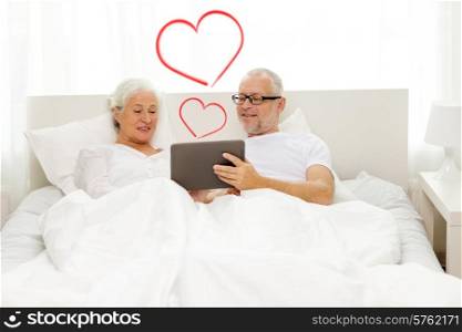 family, technology, love, age and people concept - happy senior couple looking to tablet pc computer and lying in bad at home with red heart shapes