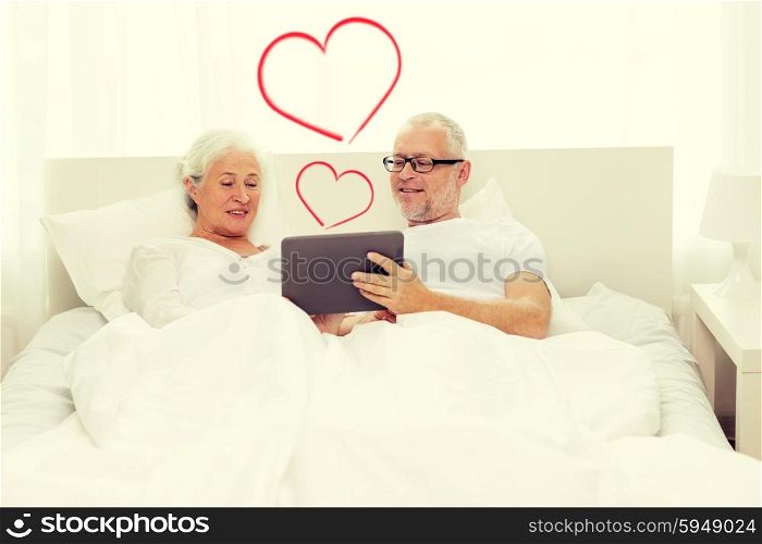 family, technology, love, age and people concept - happy senior couple looking to tablet pc computer and lying in bad at home with red heart shapes