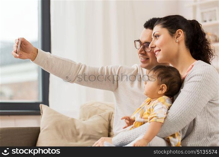 family, technology, parenthood and people concept - happy mother and father with baby daughter taking selfie by smartphone at home. mother and father with baby taking selfie at home. mother and father with baby taking selfie at home