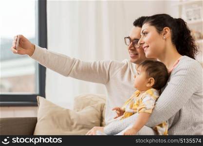family, technology, parenthood and people concept - happy mother and father with baby daughter taking selfie by smartphone at home. mother and father with baby taking selfie at home. mother and father with baby taking selfie at home
