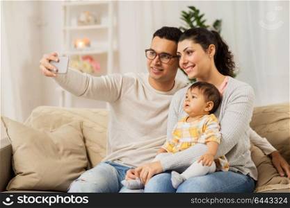 family, technology, parenthood and people concept - happy mother and father with baby daughter taking selfie by smartphone at home. mother and father with baby taking selfie at home