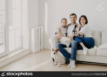 Family, togetherness and relationnship concept. Happy man embraces daughter and wife, sit on comfortable white sofa in empty room, their pet sits on floor, make family portrait for long memory