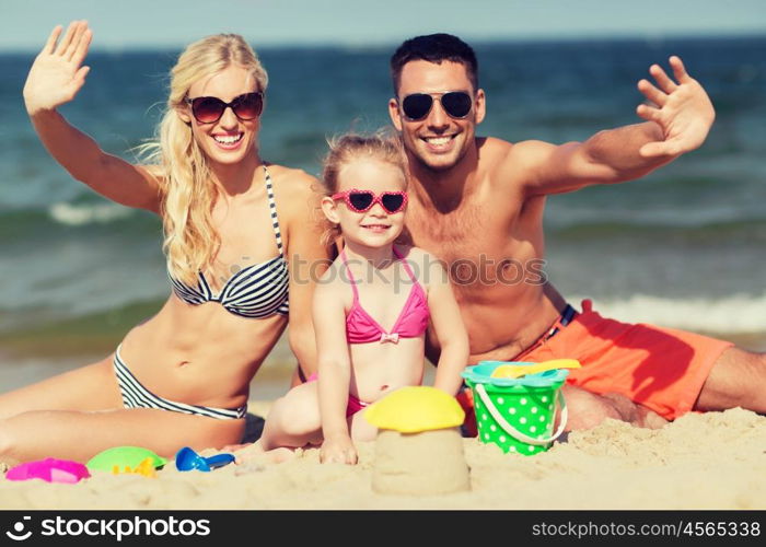 family, travel, vacation and people concept - happy man, woman and little girl in sunglasses with sand toys waving hands on summer beach