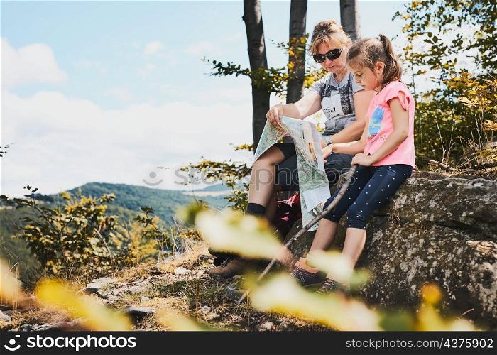 Family trip in mountains. Mother and her daughter examining a map of mountains trials sitting on rock enjoying summer day during vacation trip in mountains. People actively spending time outside