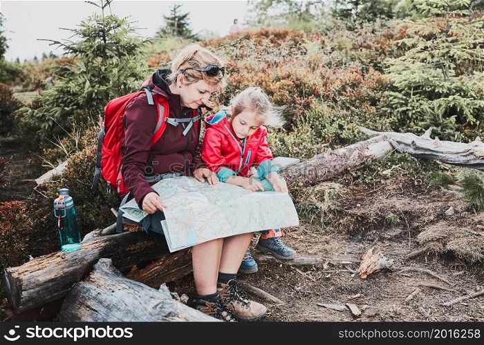 Family trip in mountains. Mother and her little daughter examining a map, sitting on stump, taking break during summer trip