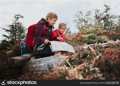 Family trip in mountains. Mother and her little daughter examining a map, sitting on stump, taking break during summer trip