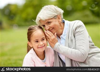family, trust and people concept - happy granddaughter whispering secrets to grandmother at summer park. granddaughter sharing secrets with grandmother
