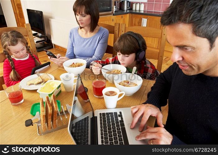 Family Using Gadgets Whilst Eating Breakfast Together In Kitchen