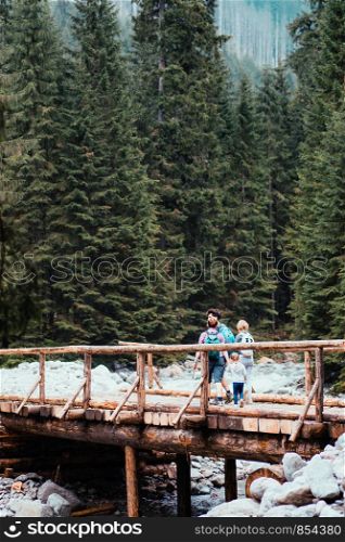 Family walking across a wooden bridge over a mountain stream. Spending vacation on wandering with backpacks in a mountains and forests
