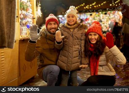 family, winter holidays and celebration concept - happy mother, father and little daughter waving hands at christmas market on town hall square in tallinn, estonia. happy family waving hands at christmas market