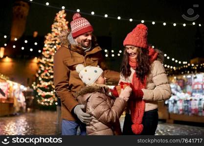 family, winter holidays and celebration concept - happy mother, father and little daughter with gift at christmas market on town hall square in tallinn, estonia. happy family with gift at christmas market in city