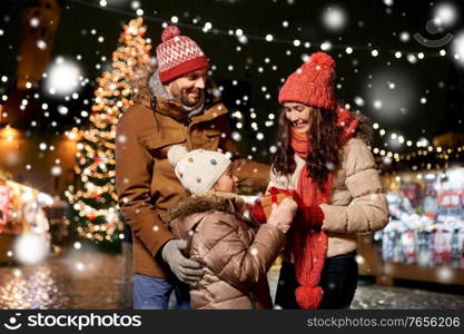 family, winter holidays and celebration concept - happy mother, father and little daughter with gift at christmas market on town hall square in tallinn, estonia over snow. happy family with gift at christmas market in city