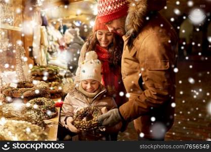 family, winter holidays and celebration concept - happy mother, father and little daughter buying wreath at christmas market on town hall square in tallinn, estonia over snow. happy family buying wreath at christmas market