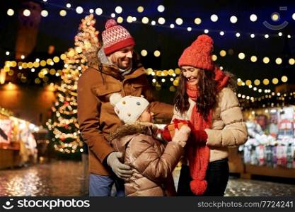 family, winter holidays and celebration concept - happy mother, father and little daughter with gift at christmas market on town hall square in tallinn, estonia. happy family with gift at christmas market in city