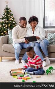 family, winter holidays and people concept - happy african american mother and father using tablet pc computer and baby son playing with toy blocks and dinosaur at home on christmas. happy african american family on christmas at home